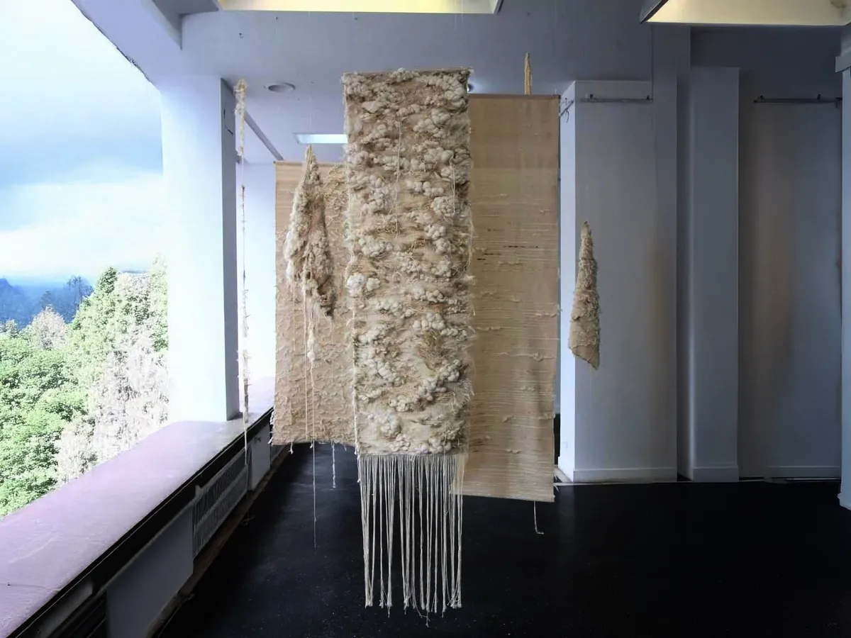 Tall cream panels suspended in a gallery that opens onto a forest; the nearest is thick with tufts of raw wool, its warp falling in a long fringe to the floor.