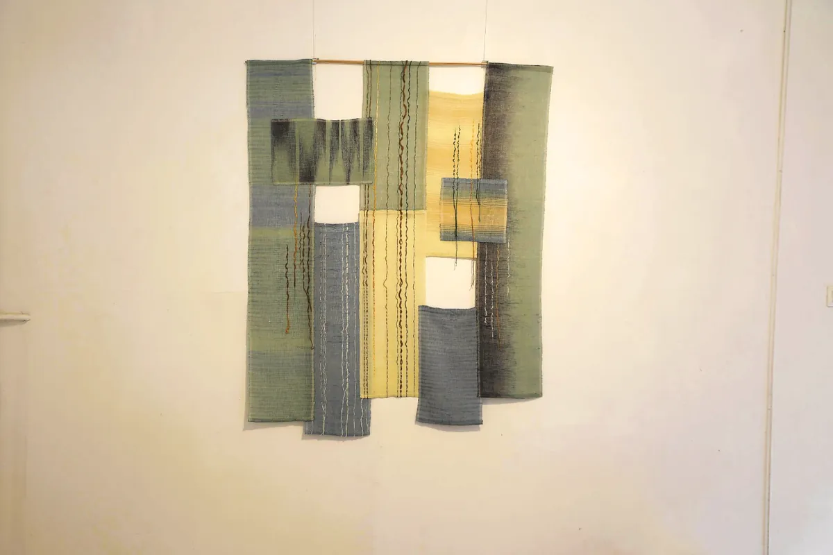 Wall piece of overlapping woven strips in sage green, slate blue and pale yellow, with loose threads falling vertically across them.