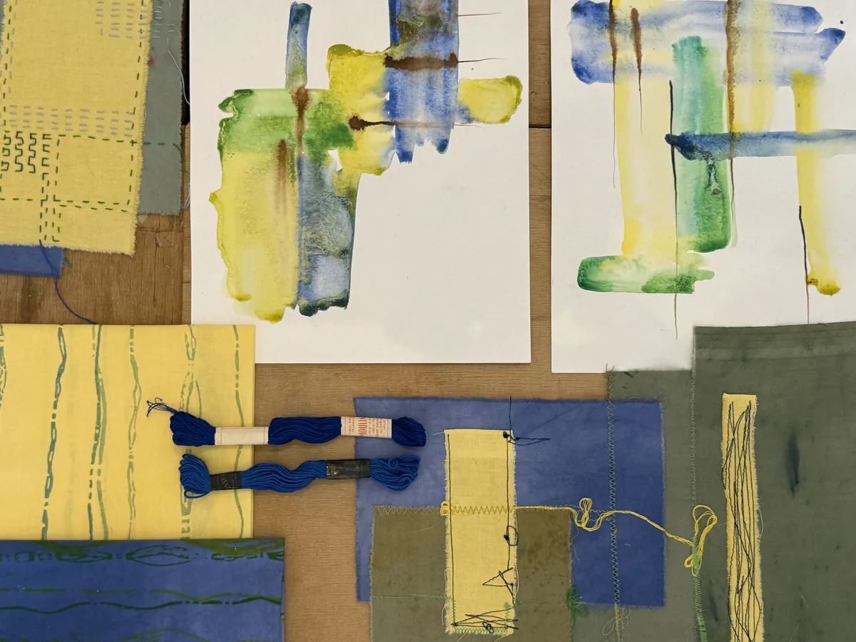 Watercolour studies in yellow, blue and green beside dyed fabrics, stitched patches and skeins of blue embroidery thread.